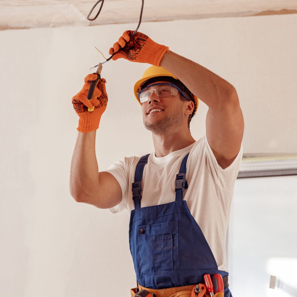 Handsome young man in safety helmet using pliers and smiling while fixing electrical wiring